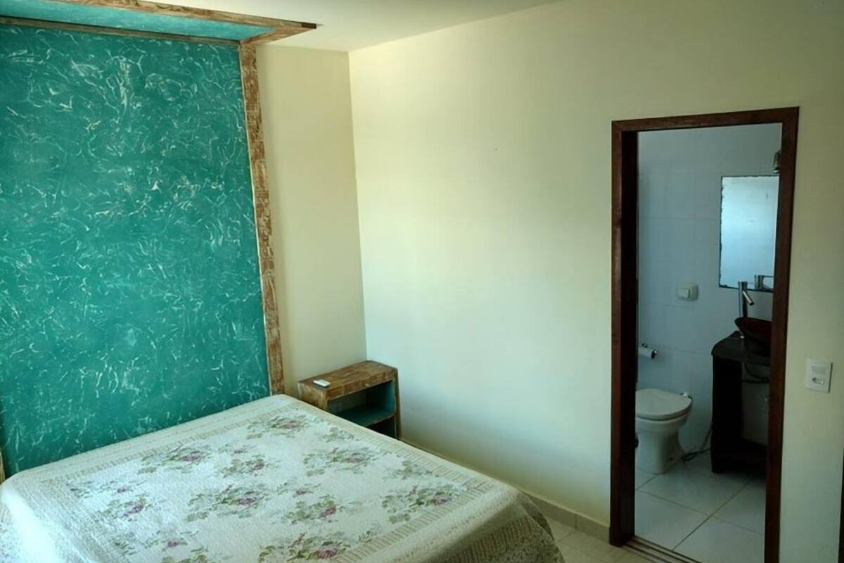 A bedroom with a bed, a small wooden nightstand, a teal-colored wall, and a doorway leading to a bathroom with a toilet and a mirror.