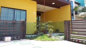 Um edifício moderno com fachada amarela, uma varanda com guarda-corrente e um pequeno jardim com palmeira e plantas em vasos.
