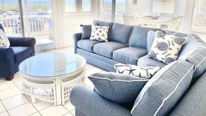 Ein geräumiger Wohnbereich mit einem blauen Sofa, einem runden Glastisch und Meerblick durch große Fenster.