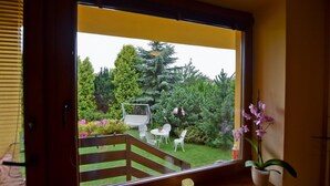 Una ventana con vista a un jardín, que incluye una terraza de madera, sillas blancas y una planta en maceta.