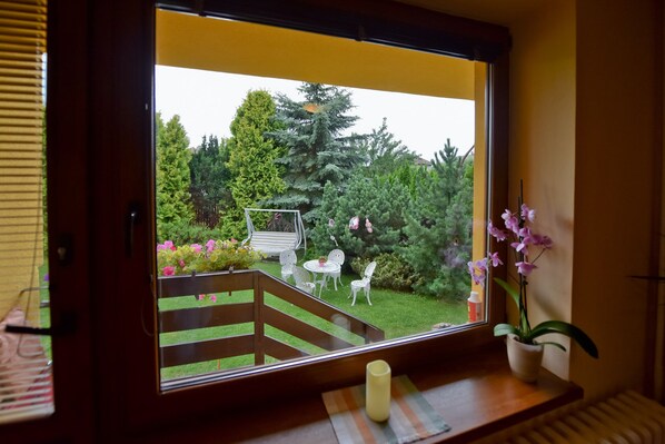 Une fenêtre donnant sur un jardin, avec une terrasse en bois, des chaises blanches et un pot de fleurs.