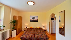 Un dormitorio con una cama doble, dos mesitas de noche con lámparas, un radiador, un armario de madera, un refrigerador y un cuadro colgado en la pared.