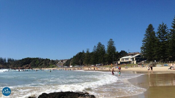 A beach with people swimming and walking along the shore. There are buildings and trees in the background. - Salt by the sea unit 3 (Port Macquarie)