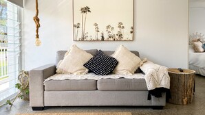 A modern living room with a grey sofa, a large framed artwork of palm trees, and a hanging light fixture. - Salt by the sea unit 3 (Port Macquarie)