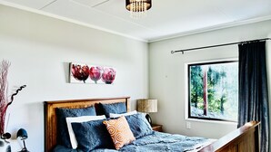 Ein Schlafzimmer mit Holzkopfteil, einem großen Bett mit blauer Bettwäsche, einer Nachttischlampe, einem Fenster mit Blick auf Bäume und einem Wandbild mit Blumen.