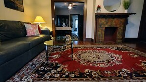 Un salon avec un canapé en cuir foncé, une table basse en verre, un tapis à motifs rouges, une cheminée et, au fond, un coin repas.
