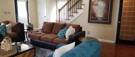 A living room with a brown sectional sofa, a blue armchair, and a coffee table. There is a staircase with a wooden railing and a painting on the wall.