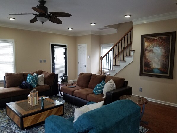 A living room with a brown sofa set, a glass coffee table, and a blue armchair. There is a painting on the wall and a staircase in the background.
