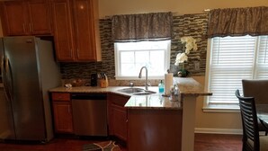 Una cocina con gabinetes de madera, un refrigerador de acero inoxidable, un lavavajillas, una encimera de granito, una ventana con cortina y un comedor con una silla.