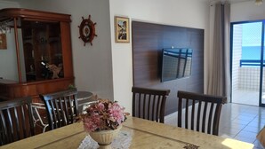 A dining area with a wooden table, chairs, a vase of flowers, a wooden cabinet, a wall-mounted TV, and a view of the outside through a glass door.