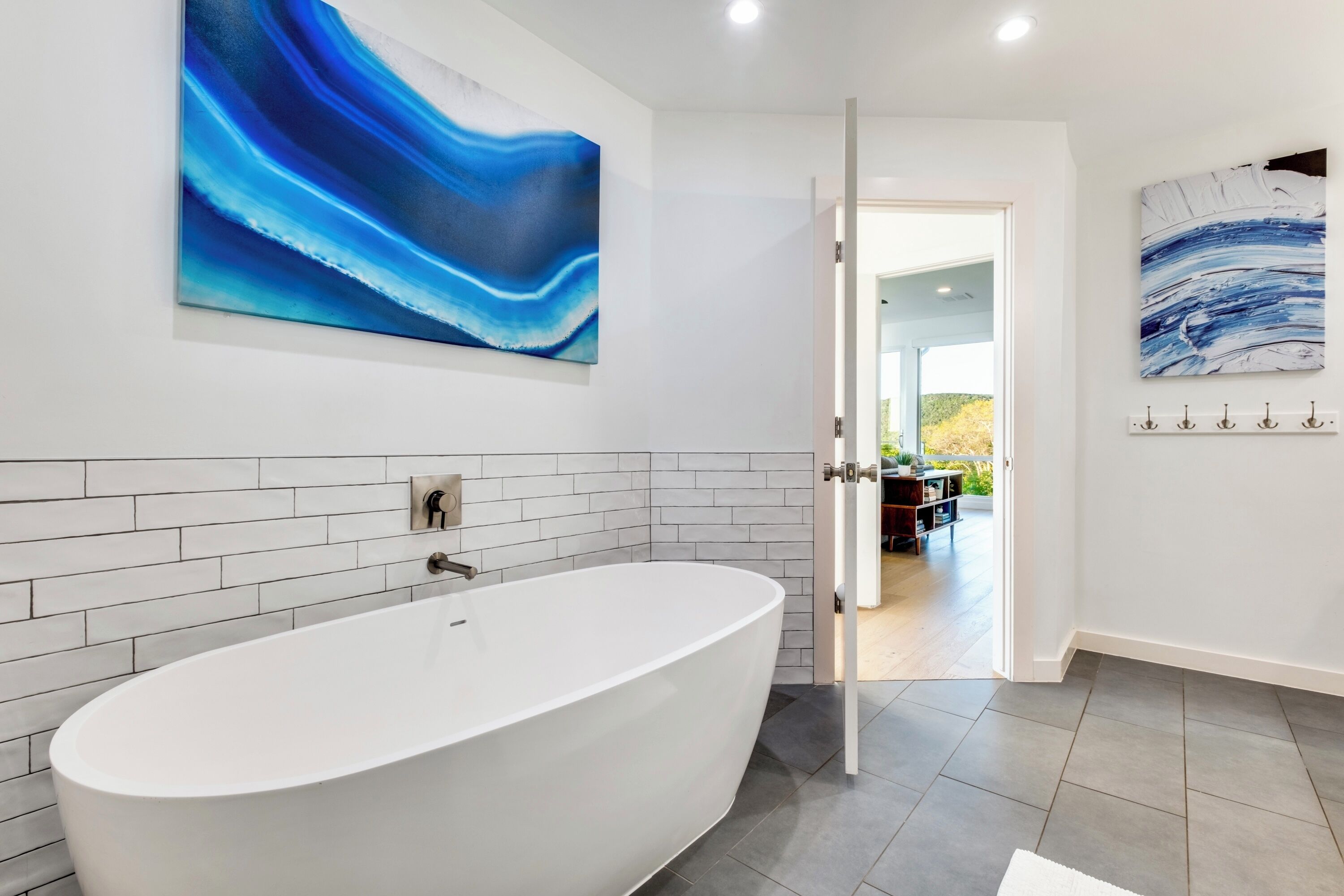 A modern bathroom with a freestanding bathtub, white tiled walls, and two abstract paintings.