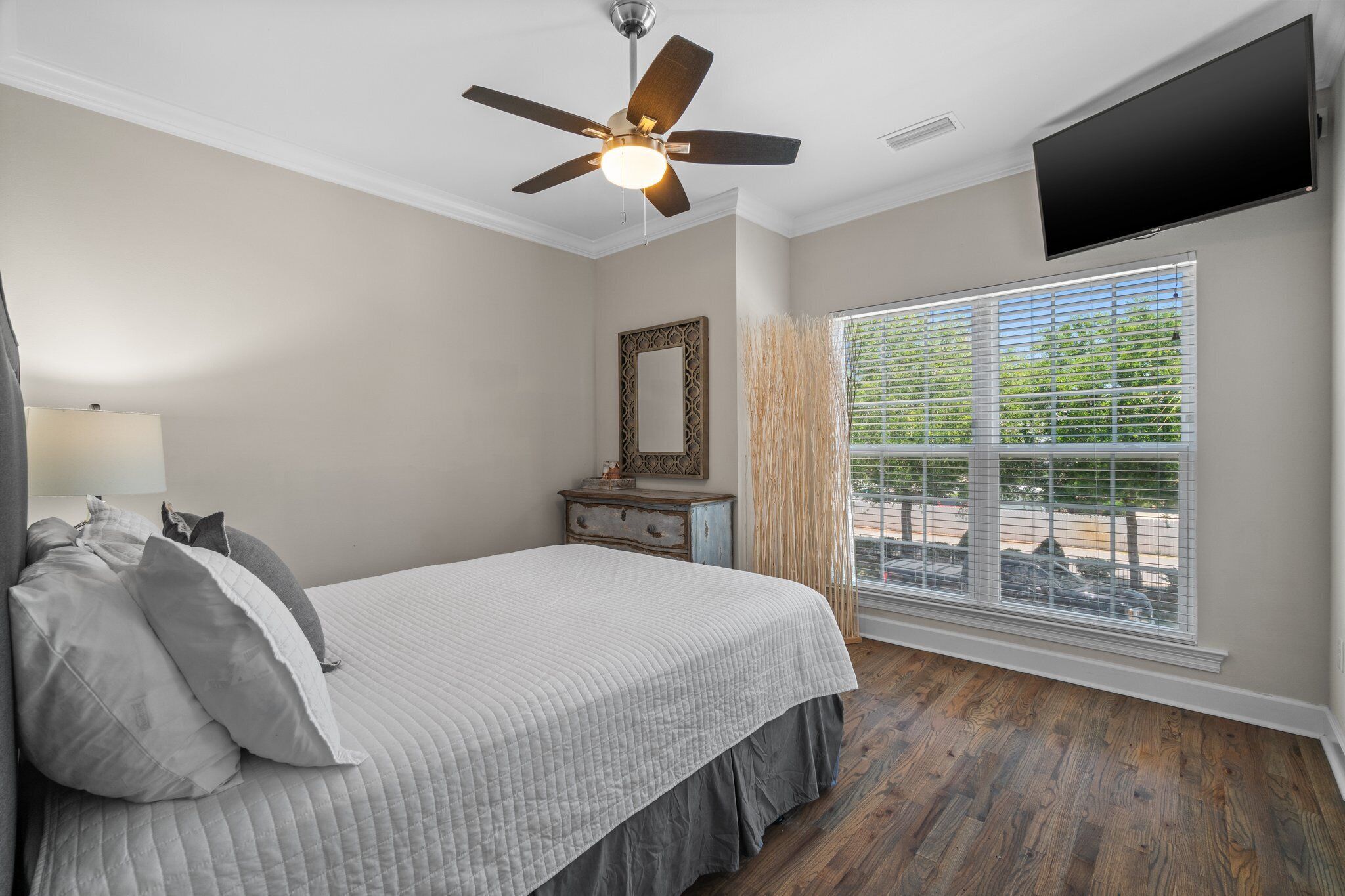 A bedroom with a bed, a ceiling fan, a TV mounted on the wall, and a window with blinds.