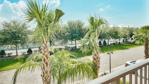 Un balcón con vistas a una calle pavimentada, palmeras y un área verde con una cerca y árboles al fondo.