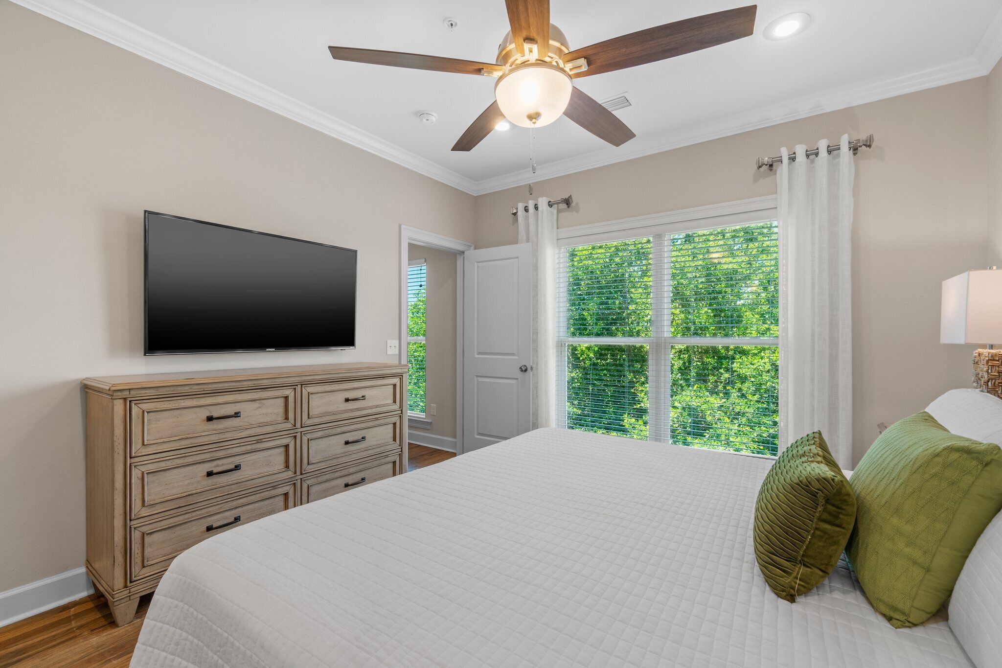 A bedroom with a bed, a dresser, a ceiling fan, a TV, and a window with curtains.