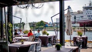 Un área de comedor al aire libre con mesas y sillas, con vista a un cuerpo de agua y un crucero al fondo.