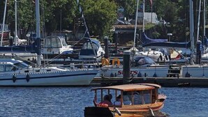 Una marina con barcos, un barco de madera con pasajeros y un edificio moderno con techo de cristal al fondo.
