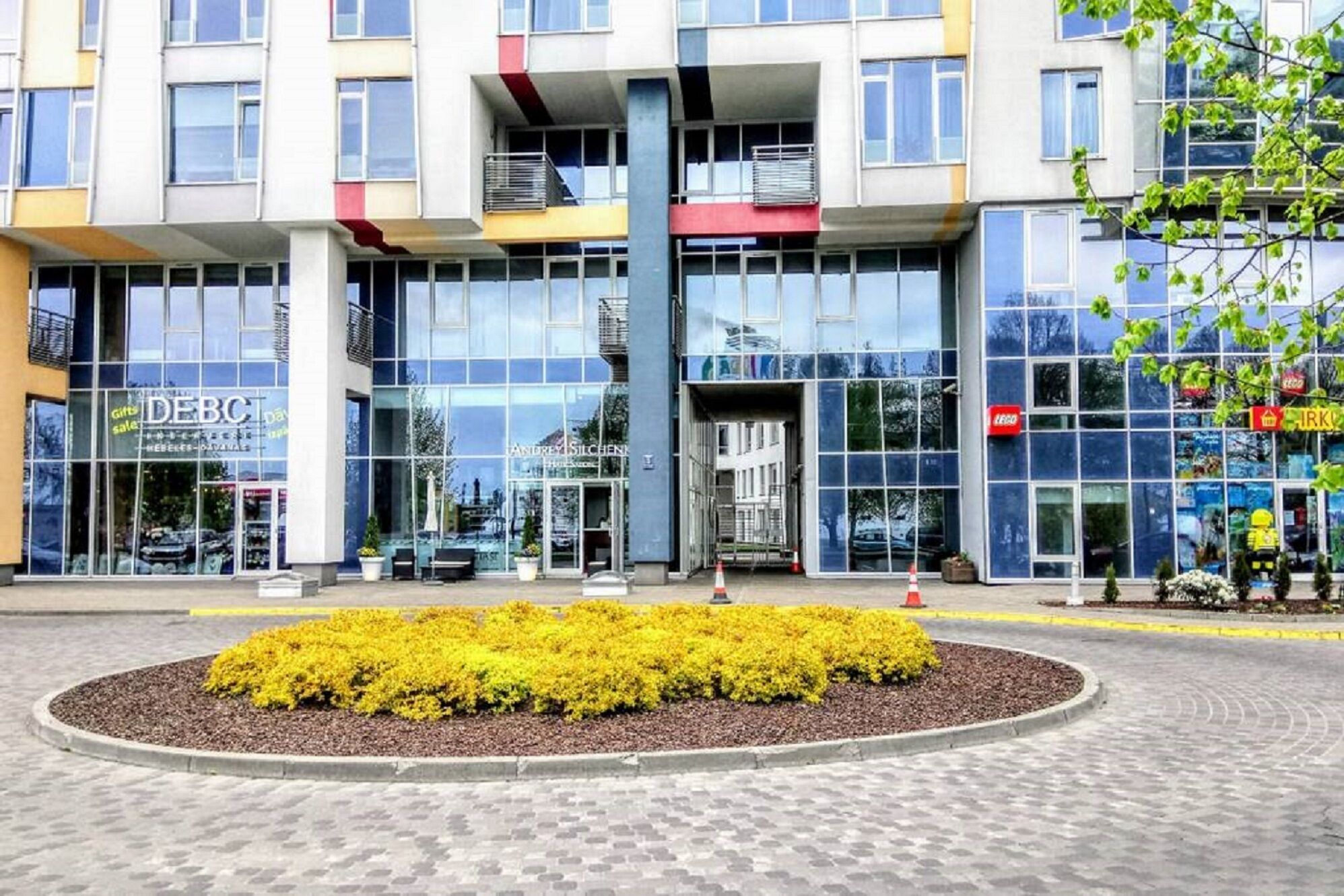 A modern building with large windows, a central entrance, and a landscaped area with yellow shrubs.