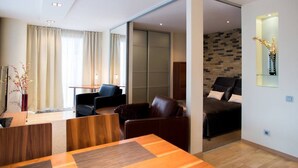 Habitación de hotel moderna con una mesa de comedor de madera, dos sofás de cuero, una pequeña cocina y un amplio ventanal corredero que da a un balcón.