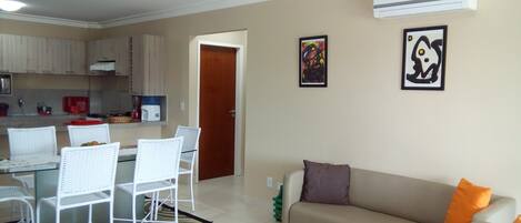 A living room with a beige sofa, a dining area with white chairs, a kitchen with wooden cabinets, and two framed pictures on the wall.