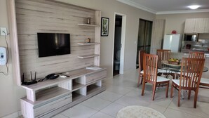 Un salon moderne doté d’un téléviseur à écran plat, d’un meuble de rangement en bois, d’une table à manger avec des chaises, et d’un coin cuisine à l’arrière-plan.
