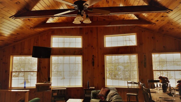 A wooden cabin interior with a ceiling fan, large windows, a television, a sofa, a dining table set for two, and a bookshelf. - BLOW OUT-SALE PRICES!  3 DAY FOLIAGE,Fishing, Dock, Fire Pit, Boat launch & Boat (North Hero)