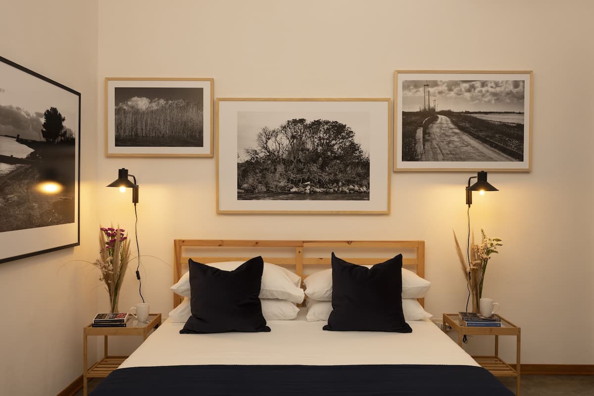 A neatly arranged bedroom with a wooden headboard, a bed with black and white bedding, two bedside tables with lamps, and three framed pictures on the wall.