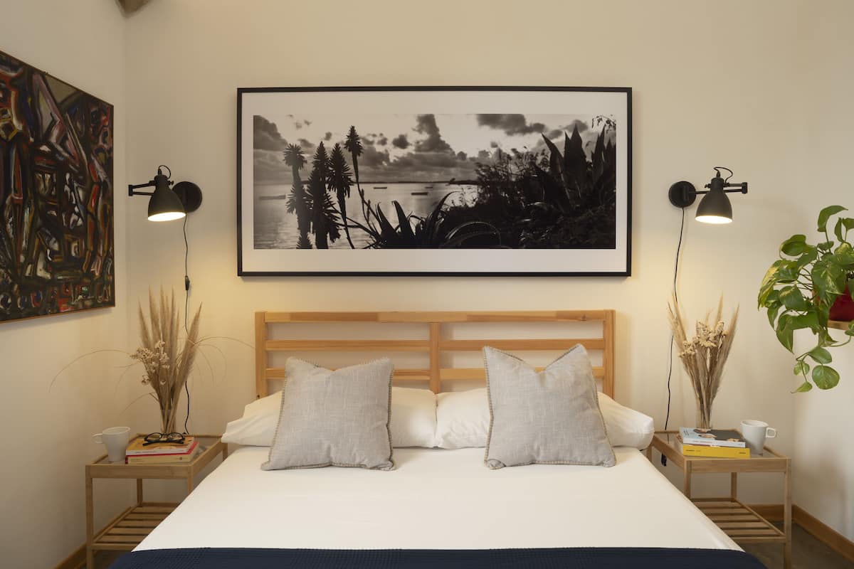 A bedroom with a large bed, two bedside tables, a headboard, a framed picture on the wall, and a potted plant.