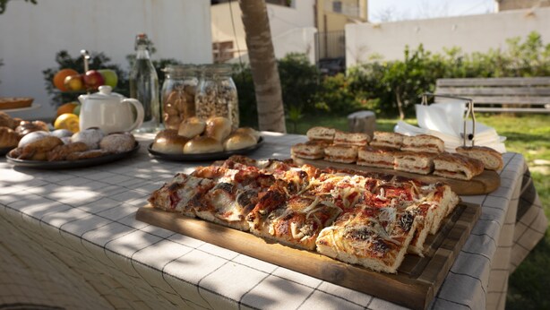 A table with various food items including pizza, bread, and pastries, set outdoors with a tree and buildings in the background. - Stagnone Area Zero (Marsala)