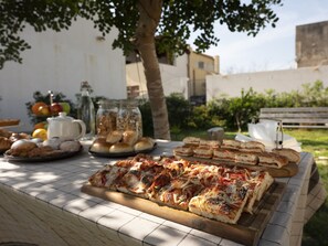 A table with various food items including pizza, bread, and pastries, set outdoors with a tree and buildings in the background. - Stagnone Area Zero (Marsala)