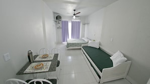 A small, clean room with a bed, a table with a chair, and a ceiling fan. - Residencial Recanto dos Pássaros (Itapema)
