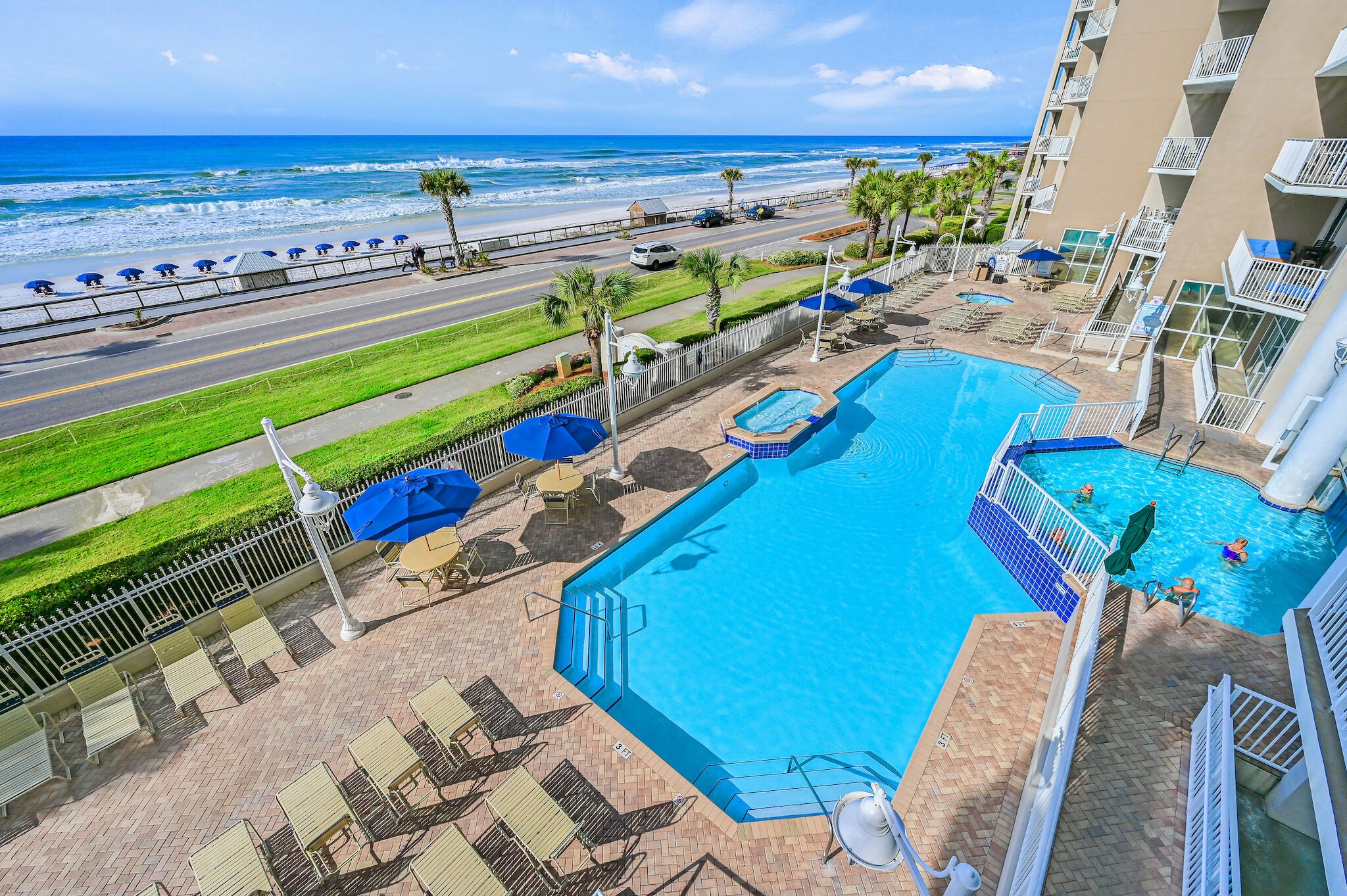 A hotel pool area with a view of the beach and ocean, a road with cars, and a fenced walkway.