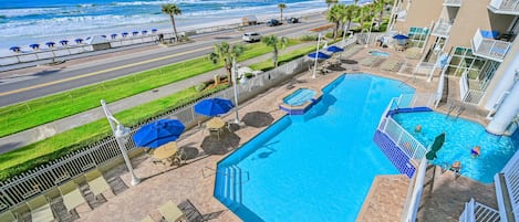 A hotel pool area with a view of the beach and ocean, a road with cars, and a fenced walkway.