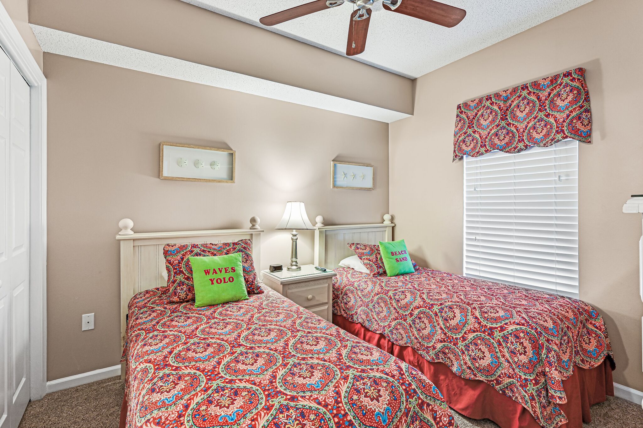 Un dormitorio con dos camas, ventilador de techo, ventana con persianas y un valance decorativo.