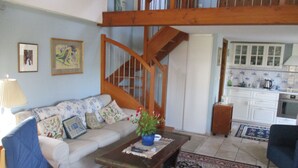 Un salon avec un escalier en bois, un canapé agrémenté de coussins à motifs floraux, une table basse avec une plante, et un coin cuisine avec des placards et un dosseret carrelé.
