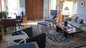 Un salon comprenant un canapé, un fauteuil, une table basse et une bibliothèque. On y trouve une porte en bois et une fenêtre avec des rideaux.