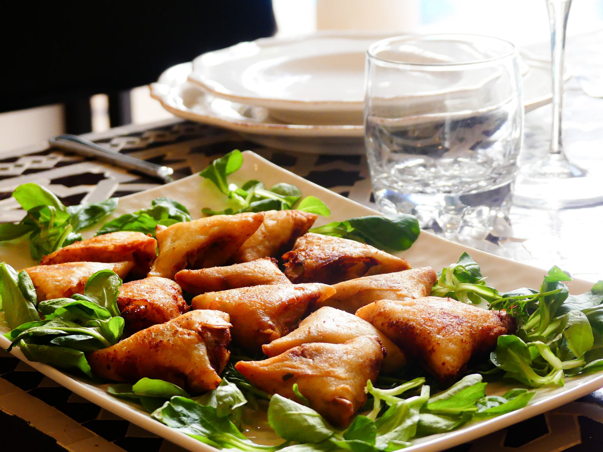 Un plato de samosas adornadas con vegetales, acompañado de un vaso de agua y un plato blanco al fondo.