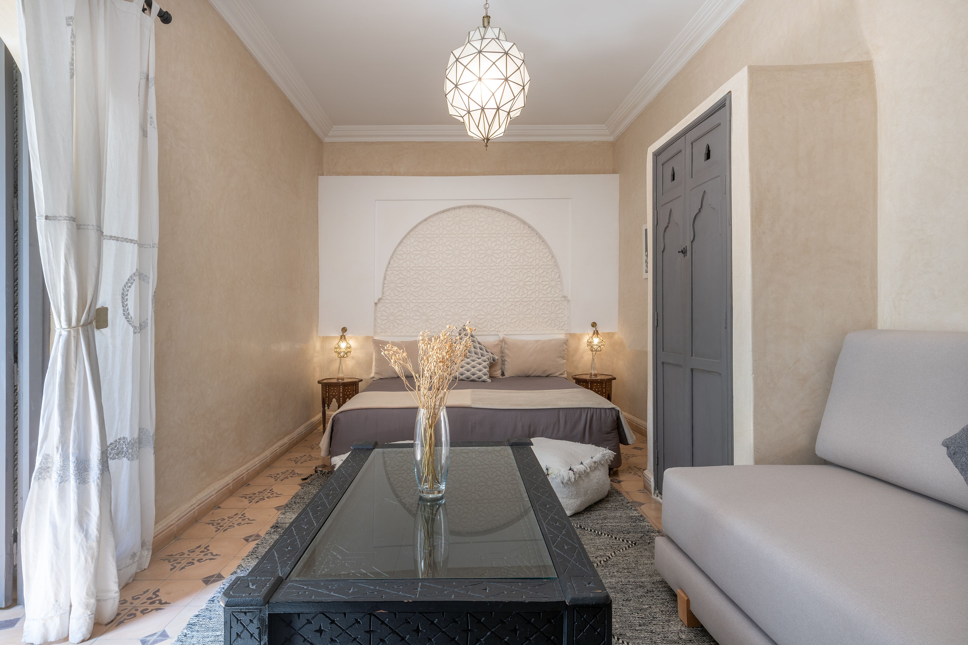 A bedroom with a bed, bedside tables, a glass-top table, a sofa, and a decorative ceiling light.