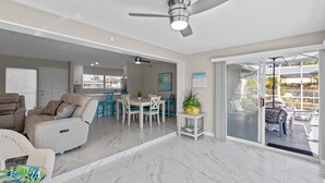 Una sala de estar moderna con sofá, mesa de comedor, sillas, una zona de cocina y un gran ventanal corredera que da a un patio exterior.