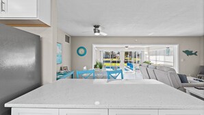 Una cocina moderna con una isla grande, un comedor con sillas azules y vistas a una piscina a través de puertas de cristal.