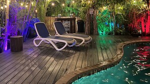 Une terrasse en bois avec deux chaises longues et une petite table, entourée d’une végétation luxuriante et d’une piscine.