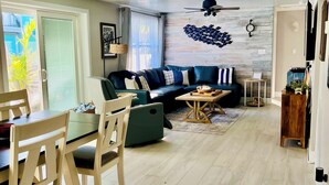 Un salon moderne avec un canapé, une table à manger et des chaises. On y trouve un ventilateur de plafond, un téléviseur fixé au mur et une fenêtre avec des stores.