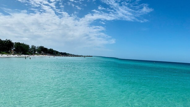 Une plage à l’eau turquoise et limpide, un rivage sablonneux et, en arrière-plan, une colline boisée.