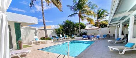 Un área de piscina con sillones de descanso, palmeras y un cielo azul despejado.