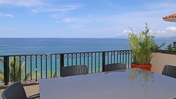 A balcony with a view of the sea, a table, chairs, and a potted plant.