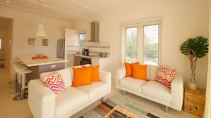 A modern kitchen with white cabinets, a stainless steel refrigerator, and a dining area with white chairs. There are two white sofas with orange pillows, a large plant, and a ceiling fan. - Bayfront 1BR Apt - Central Location & GREAT Rates! (Governor`s Harbour)
