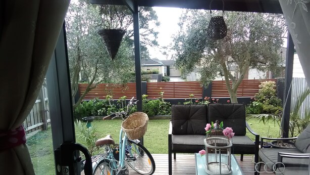 Een overdekt terras met een houten terras, een blauwe fiets met mandje, een comfortabele bank en een tafel met een vogelkooi.