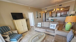 A living room with a sofa, armchairs, a television on a cabinet, and a dining area with chairs and a table. - Inn at Dauphin Island 310 Beautiful Unit with Wonderful Views!! (Dauphin Island)
