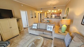 A living room with a sofa, armchairs, a coffee table, and a TV. There is a kitchen in the background. - Inn at Dauphin Island 310 Beautiful Unit with Wonderful Views!! (Dauphin Island)