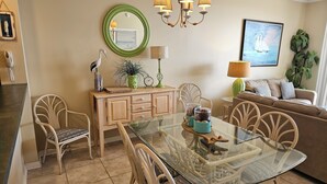 A dining area with a glass-top table, wicker chairs, a wooden cabinet, and a green-framed mirror. - Inn at Dauphin Island 310 Beautiful Unit with Wonderful Views!! (Dauphin Island)