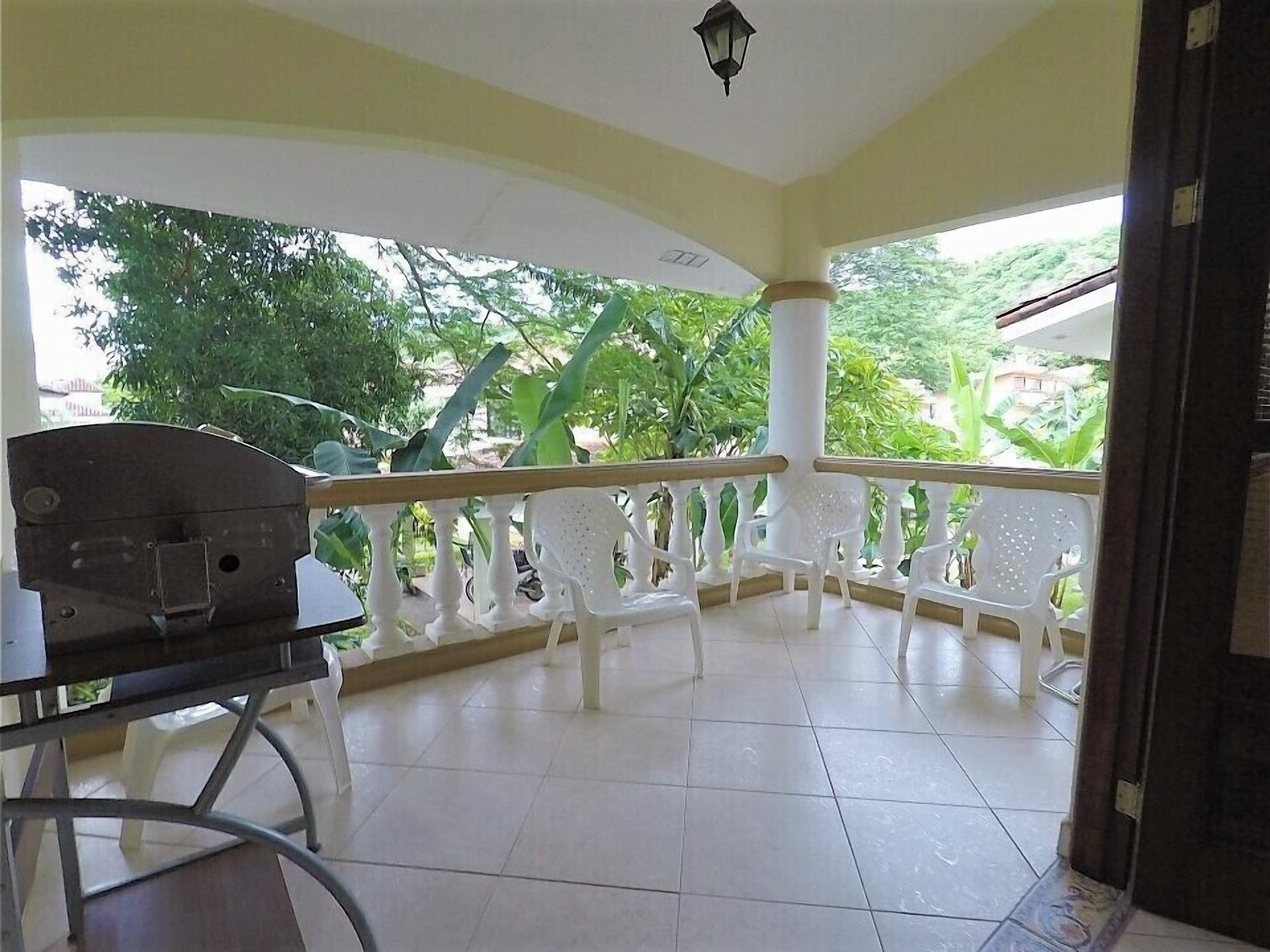 A covered balcony with white plastic chairs, a table, and a view of greenery outside.